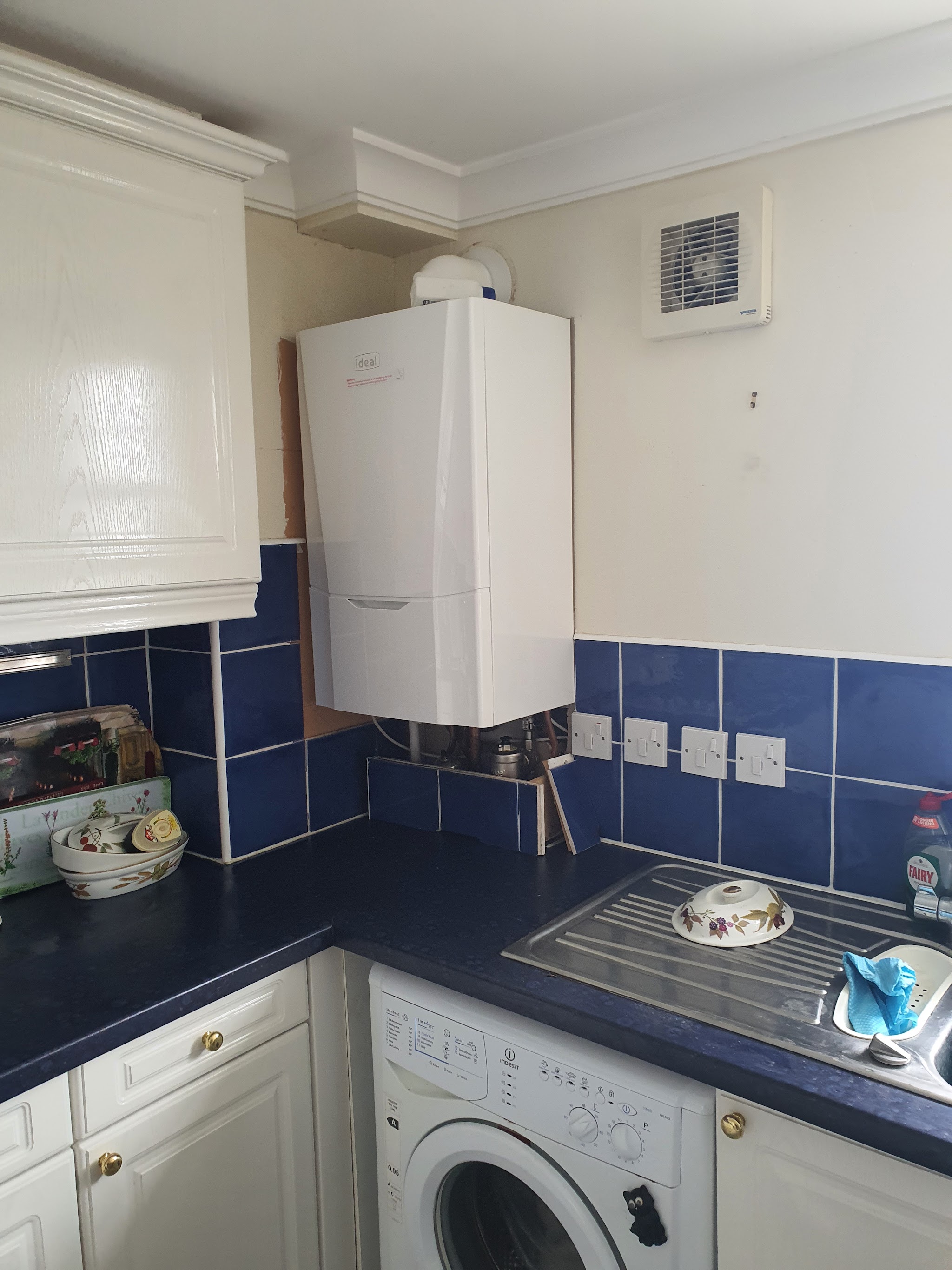 A white boiler fitted in a kitchen corner above a blue-tiled worktop and washing machine